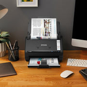 An Epson ES-550W document scanner sits on a wooden desk in a home office setup, scanning a printed page from a magazine. The desk also contains a keyboard, mouse, laptop, and other office accessories against a dark grey wall.